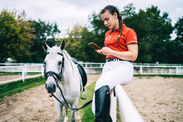 Side view of female horse rider sitting on metal fence and holding bridle in hand while looking at smartphone near horse