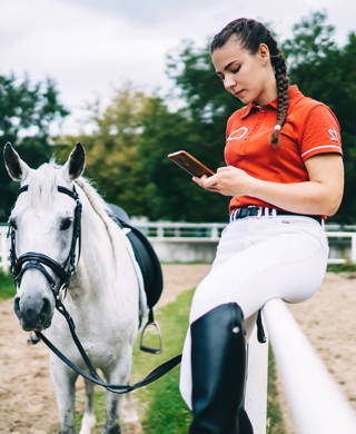 Side view of female horse rider sitting on metal fence and holding bridle in hand while looking at smartphone near horse
