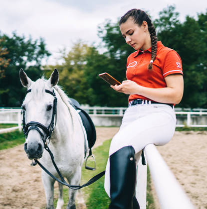 Side view of female horse rider sitting on metal fence and holding bridle in hand while looking at smartphone near horse
