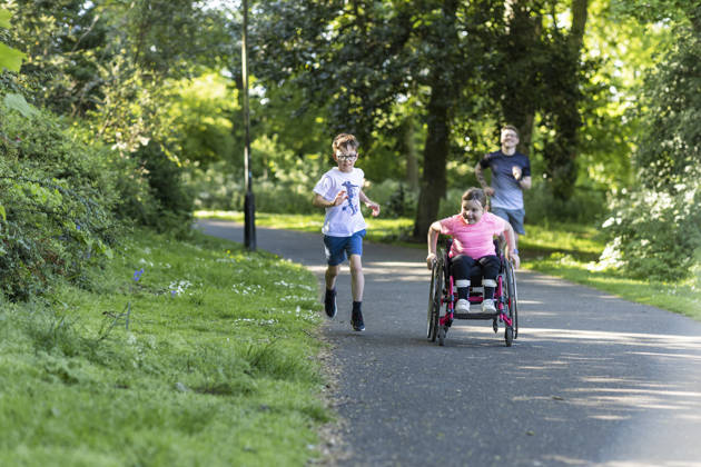 A young girl who is a wheelchair user, riding fast along a path in a public park in Newcastle Upon Tyne, England. She is being followed by her father and friend who are running after her.