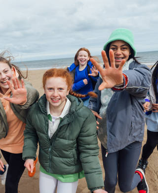 Front view of a medium group of female teenage friends wearing casual clothing on a summer day in Whitley Bay, Northeastern England. They are gathered side by side standing in a group on the beach smiling and looking at the camera with their hands raised.