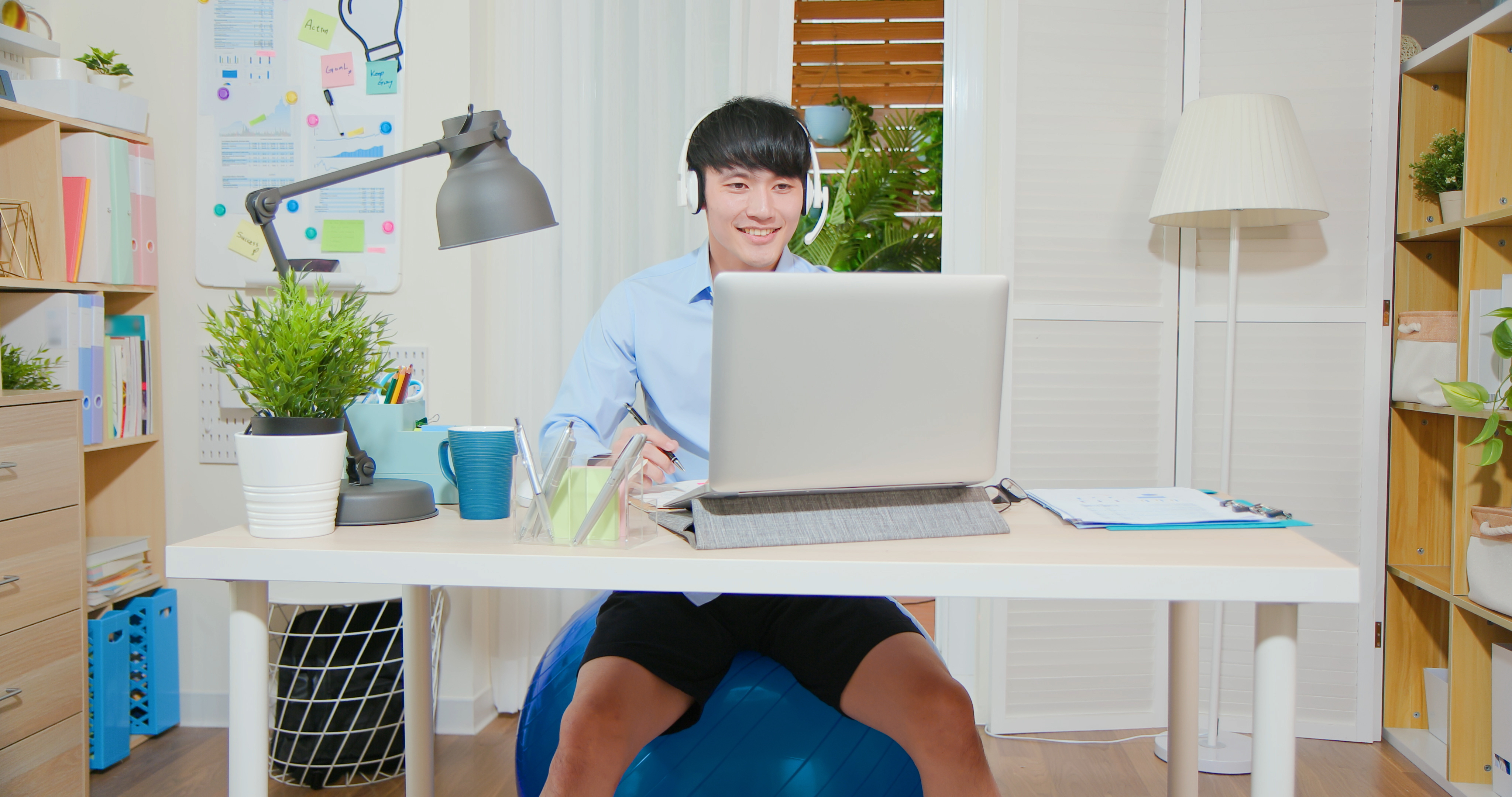 casually asian man sitting on pilates ball has video meeting - he wearing headset is using laptop  at home