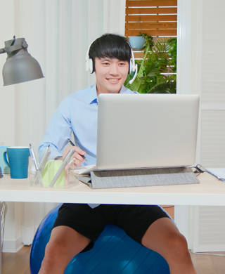 casually asian man sitting on pilates ball has video meeting - he wearing headset is using laptop at home