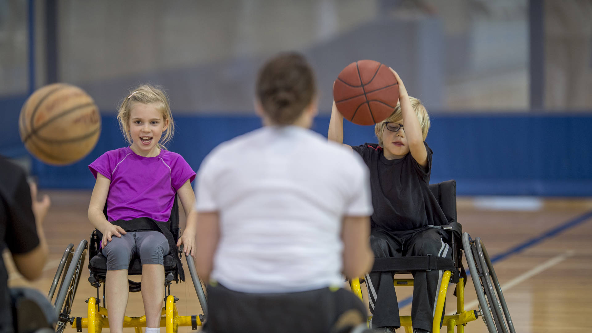 Two disabled children are bouncing a basketball in the gym with their physical therapist.