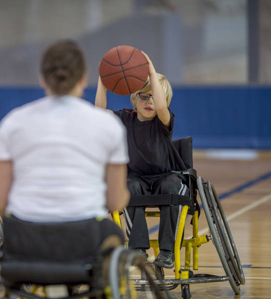 Two disabled children are bouncing a basketball in the gym with their physical therapist.