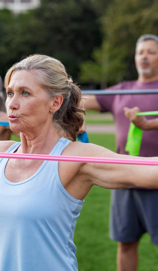 A group of older adults doing arms exercises with colorful resistance bands. Seniors standing in a park, pulling bands for strength training. Old healthy woman staying fit with stretching exercises outdoors with multiethnic friends.