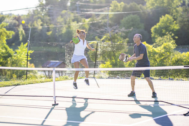 Two adults stand on one side of a net as they play Pickleball with another double.  The female is jumping to hit the ball while her partner watches in anticipation, and both are wearing comfortable athletic wear.