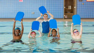 A group of children with their swimming instructor holding swimming floats above their heads in a swimming pool in Boldon, North East England. They are looking at the camera and smiling.