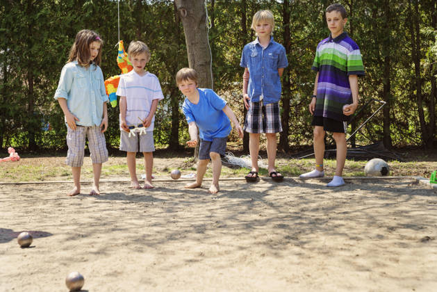 Little girl playing a ball game outdoors with the boys in a suburb backyard. She is dressed in unisex clothes, confortable for playing. They are playing petanque, a popular french game. This was taken on a hot and sunny spring day. Horizontal full length shot with copy space.