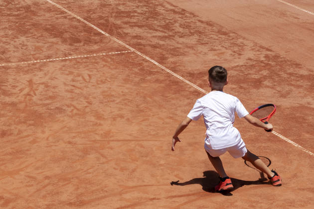 Young male tennis player with racket in action. Boy plays tennis on a clay tennis court. Child is concentrated and focused on the game. Kids tennis sport background with shadow. Motion. Copy space