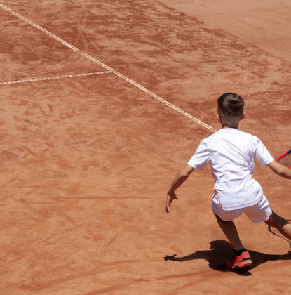 Young male tennis player with racket in action. Boy plays tennis on a clay tennis court. Child is concentrated and focused on the game. Kids tennis sport background with shadow. Motion. Copy space