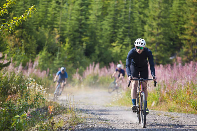 A man goes for a ride on his gravel bicycle in the Rocky Mountains of Canada. He is followed by two other riders in the background. Gravel bikes are similar to cyclo-cross bicycles with sturdy wheels and tires for riding on rough terrain. His bike has a large frame bag to carry extra gear.