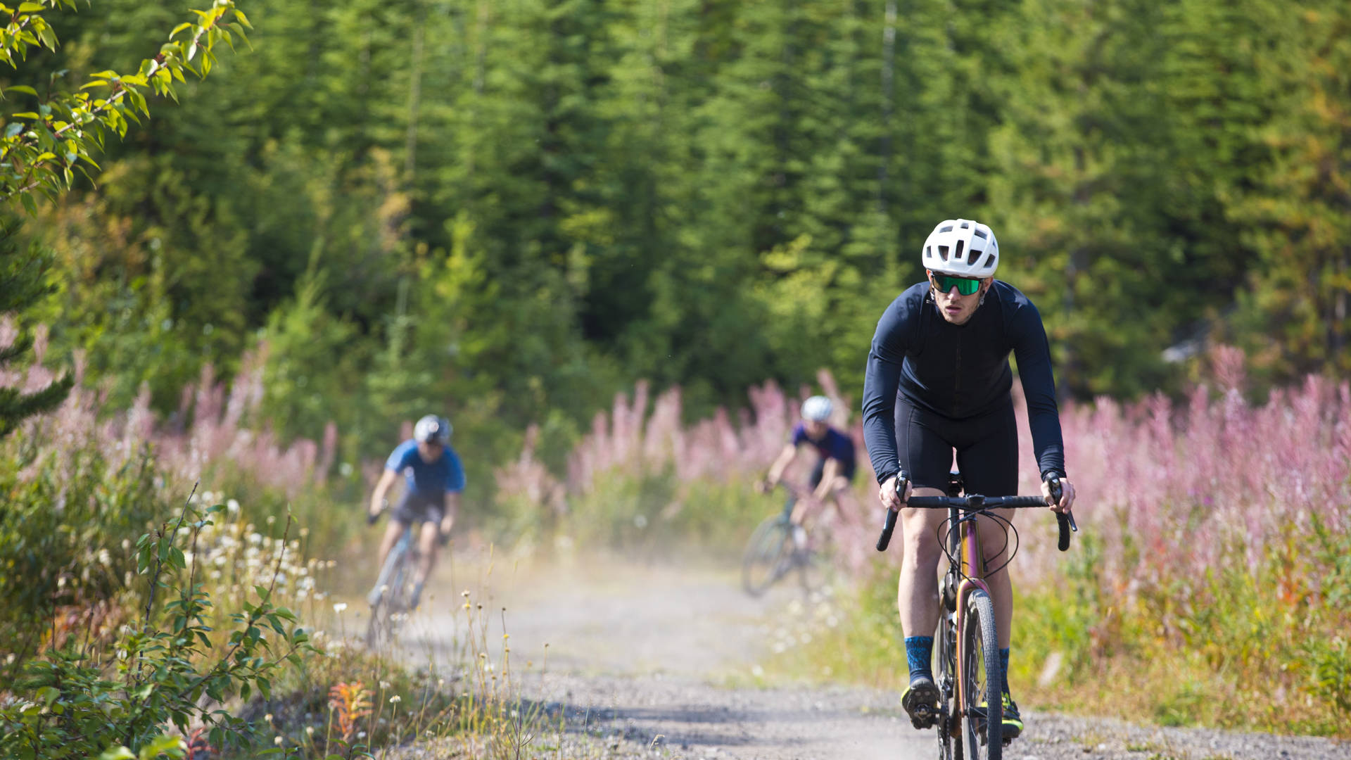 A man goes for a ride on his gravel bicycle in the Rocky Mountains of Canada. He is followed by two other riders in the background. Gravel bikes are similar to cyclo-cross bicycles with sturdy wheels and tires for riding on rough terrain. His bike has a large frame bag to carry extra gear.