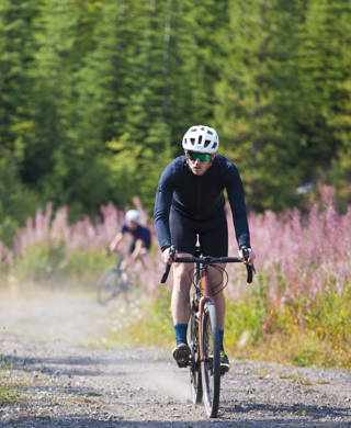A man goes for a ride on his gravel bicycle in the Rocky Mountains of Canada. He is followed by two other riders in the background. Gravel bikes are similar to cyclo-cross bicycles with sturdy wheels and tires for riding on rough terrain. His bike has a large frame bag to carry extra gear.