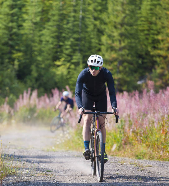 A man goes for a ride on his gravel bicycle in the Rocky Mountains of Canada. He is followed by two other riders in the background. Gravel bikes are similar to cyclo-cross bicycles with sturdy wheels and tires for riding on rough terrain. His bike has a large frame bag to carry extra gear.