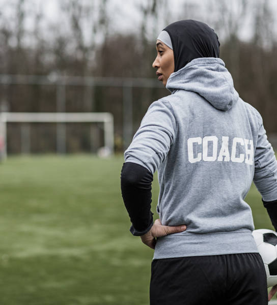 Jonge vrouwelijke voetbalcoach met hoofddoek op aan de rand van een voetbalveld