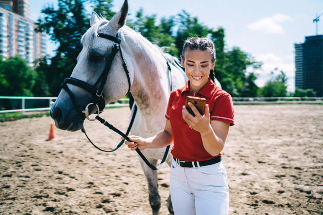 Female smiling jockey in casual outfit surfing internet on cellphone while walking grey horse on riding arena in summer afternoon