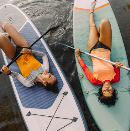 Photo of two teenage girls lying on their stand-up paddleboards and having a great time; teenagers hanging out in nature and enjoying their summer vacation.