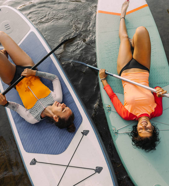 Photo of two teenage girls lying on their stand-up paddleboards and having a great time; teenagers hanging out in nature and enjoying their summer vacation.