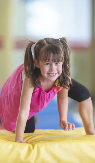 A young girl is smiling as she climbs a gymnastics landing pad. There are other children behind her. They are in a gymnasium.