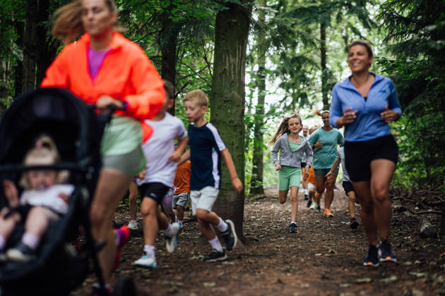 A medium shot of a group of adults and children wearing sports clothing. They are taking part in a fun run with other children and adults at Cramlington Nature Reserve in the North East of England. The focus is on a young girl as she runs with other participants.