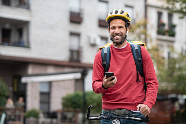Delivery food courier guy checking order via mobile phone. Smiling man with delivery bag holding smartphone and wearing helmet on cycle. Food delivery courier with yellow backpack riding bicycle on city street with copy space. Fast lunch takeaway delivery.