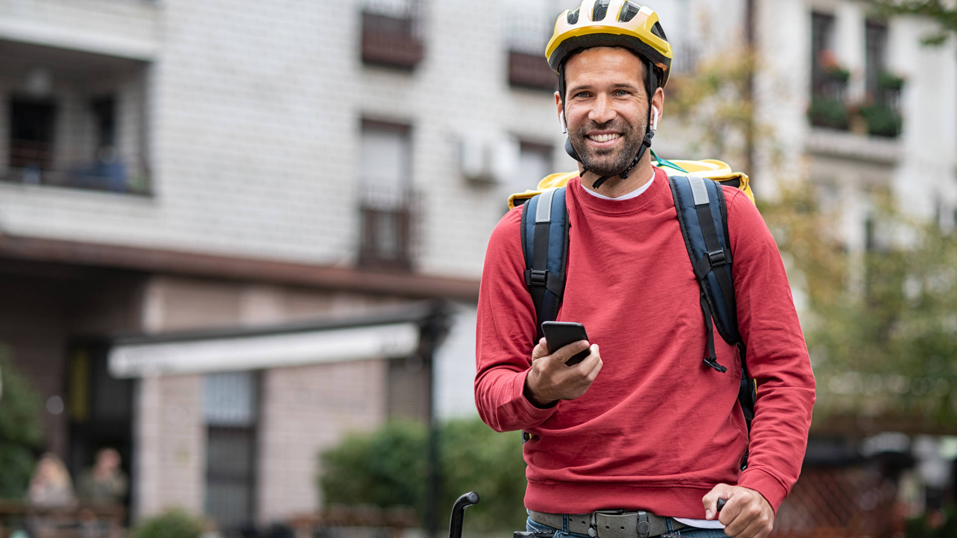 Delivery food courier guy checking order via mobile phone. Smiling man with delivery bag holding smartphone and wearing helmet on cycle. Food delivery courier with yellow backpack riding bicycle on city street with copy space. Fast lunch takeaway delivery.