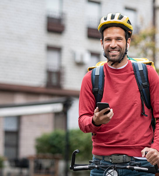 Delivery food courier guy checking order via mobile phone. Smiling man with delivery bag holding smartphone and wearing helmet on cycle. Food delivery courier with yellow backpack riding bicycle on city street with copy space. Fast lunch takeaway delivery.