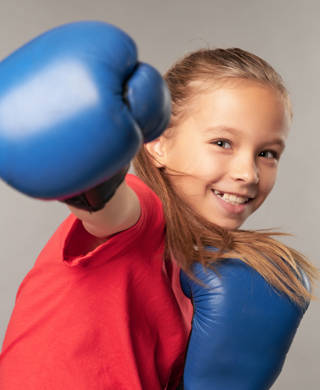 Cheerful female child in boxing gloves punching air and smiling while standing against gray background
