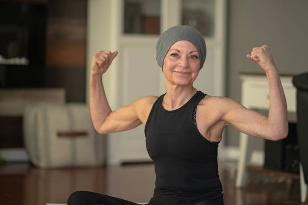 A senior woman with cancer flexes her muscles while sitting at home on her floor. She is smiling because she is getting stronger.