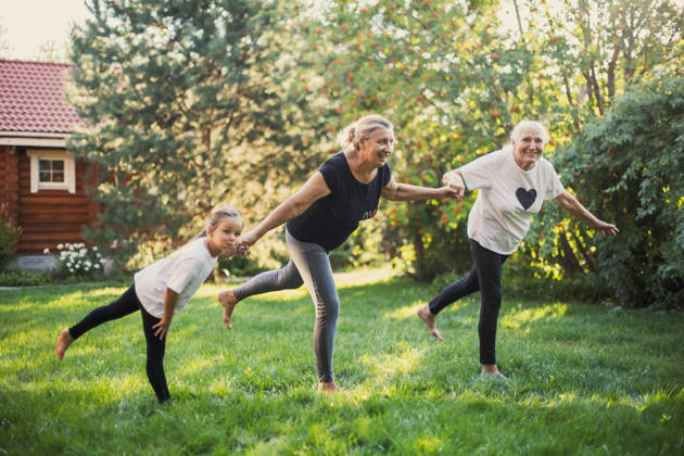 Three generations of family females smiling balancing on one leg, holding hands doing physical exercises together on backyard on meadow full of green grass and trees. Spending time together.