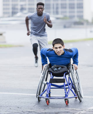 Front view of a focused young man in his 20s in sports clothing, an amputee in a self-propelled wheelchair training for a race. He is staring toward the camera with a serious expression. His African-American friend is running behind him.