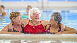A group of happy seniors taking a water aerobics class at a community centre. The women use various pool equipment to exercise.