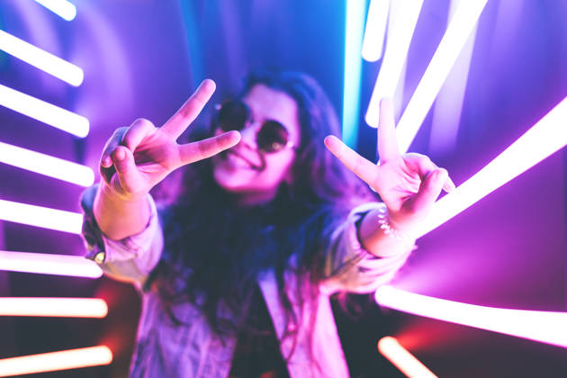 Cheerful cute smiling girl shows peace sign in round glasses at purple neon background.