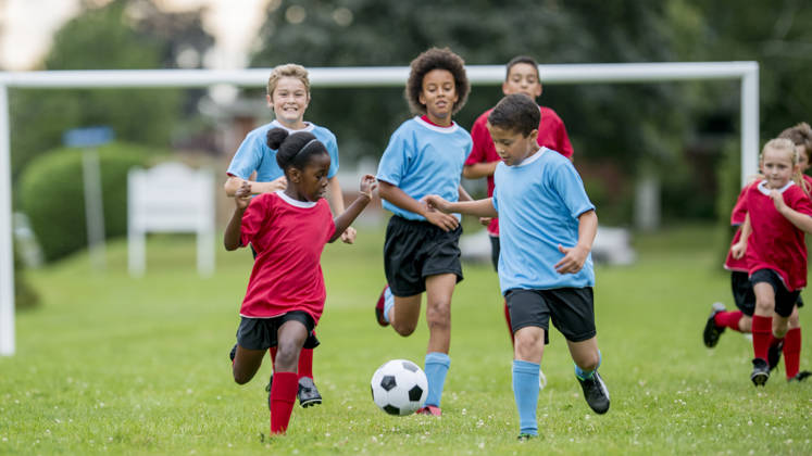 A multi-ethnic group of children are playing soccer, while running down a grass field, kicking and chasing the ball.