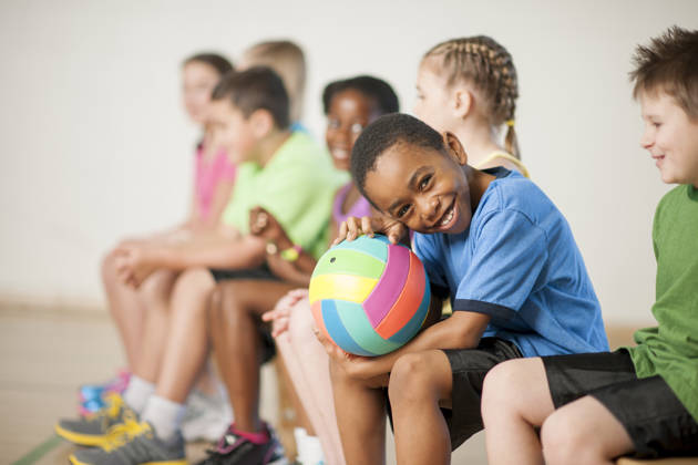 A multi-ethnic group of elementary age children are sitting in gym class and are going to play a game of volleyball. One boy is smiling while looking at the camera.