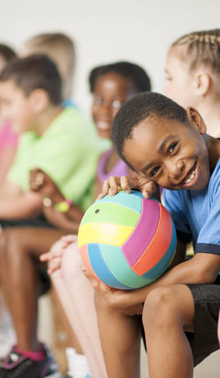 A multi-ethnic group of elementary age children are sitting in gym class and are going to play a game of volleyball. One boy is smiling while looking at the camera.