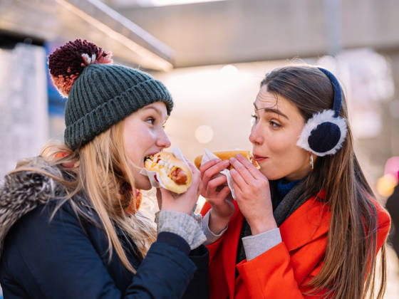 Young Caucasian teenage women eating hot dogs and smiling looking at each other while standing on the sidewalk, in downtown area of big city in North America. Both smiling, dressed in heavy winter clothes.