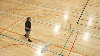 Senior man in shorts and black T-shirt cleaning the floor in a new school gymnasium, new parquet with lines and curves for different team sports(basketball, volleyball, soccer, handball). Side view from above, daylight from roof windows. Nikon D800, full frame, XXL. Motion blur.