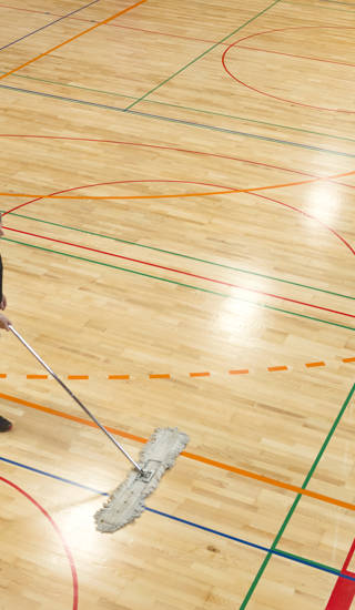 Senior man in shorts and black T-shirt cleaning the floor in a new school gymnasium, new parquet with lines and curves for different team sports(basketball, volleyball, soccer, handball). Side view from above, daylight from roof windows. Nikon D800, full frame, XXL. Motion blur.