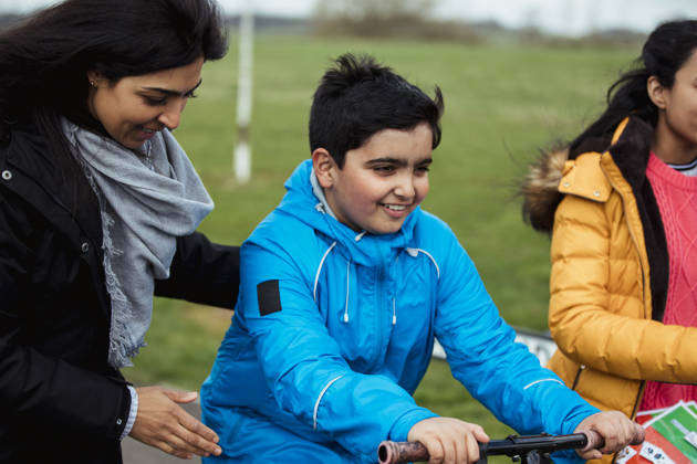 A mother is holding her son, supporting him on his scooter, with his sister walking by his side.