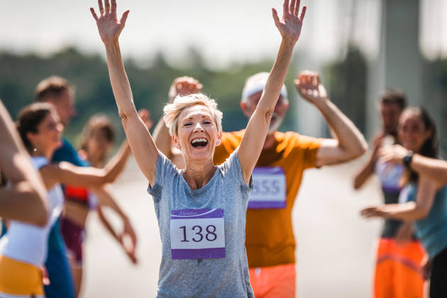 Cheerful senior woman greeting with her supporters while running a marathon on the road and looking at camera.