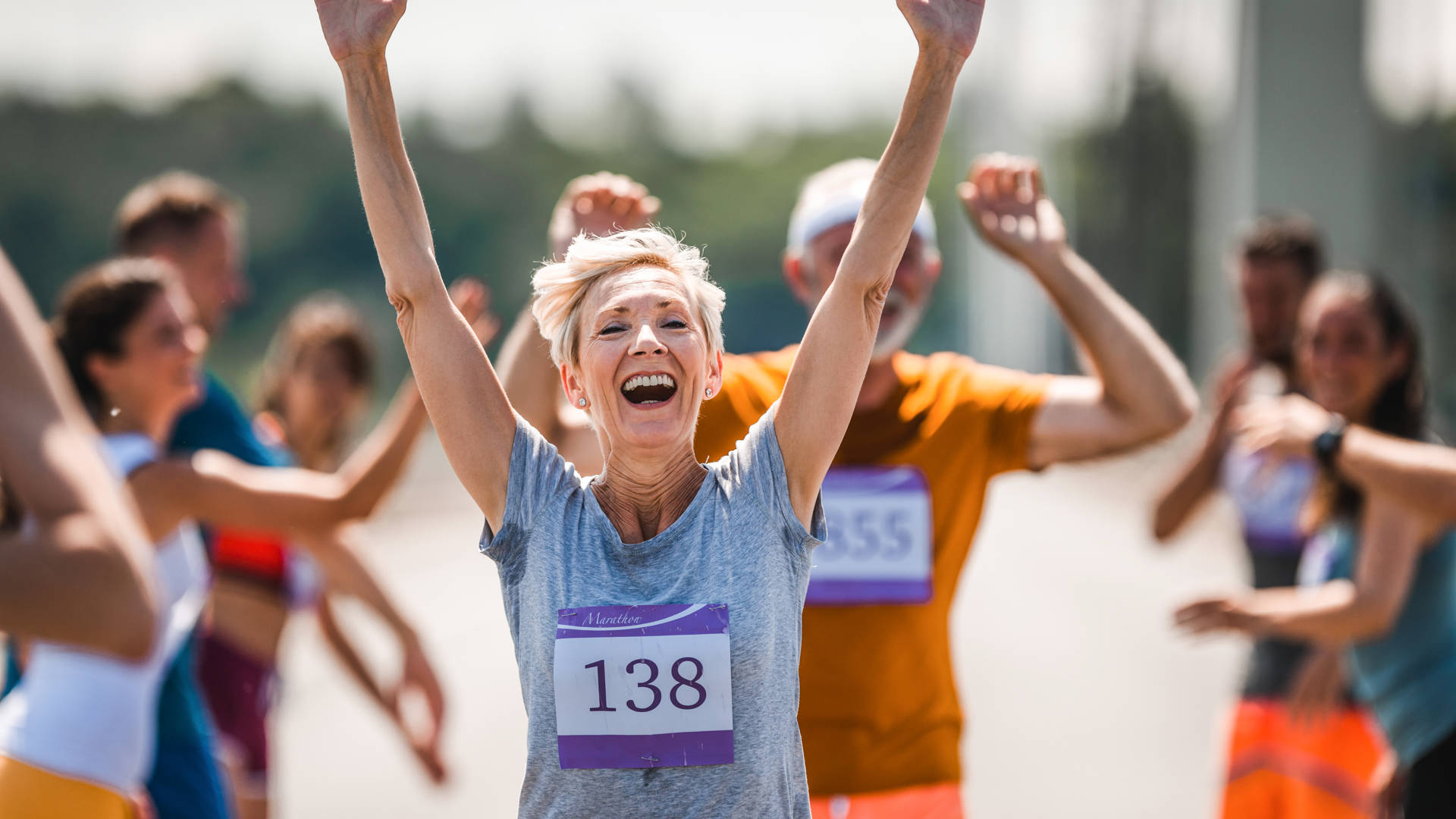 Cheerful senior woman greeting with her supporters while running a marathon on the road and looking at camera.
