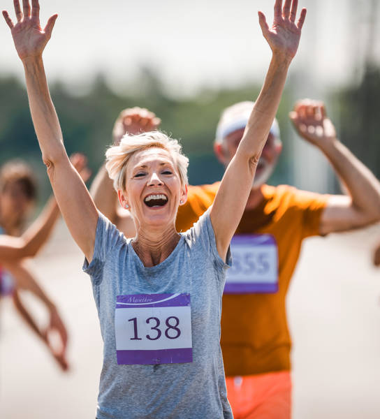 Cheerful senior woman greeting with her supporters while running a marathon on the road and looking at camera.