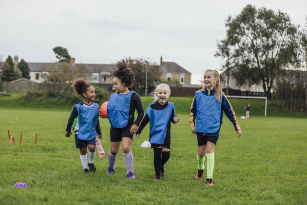 Front view of a small group of young girls wearing sports clothing, football boots and a sports bib on a football pitch in the North East of England. They are at football training where they are doing different football training drills. They are walking side by side, one girl is holding a football.
Videos are available for this scenario.