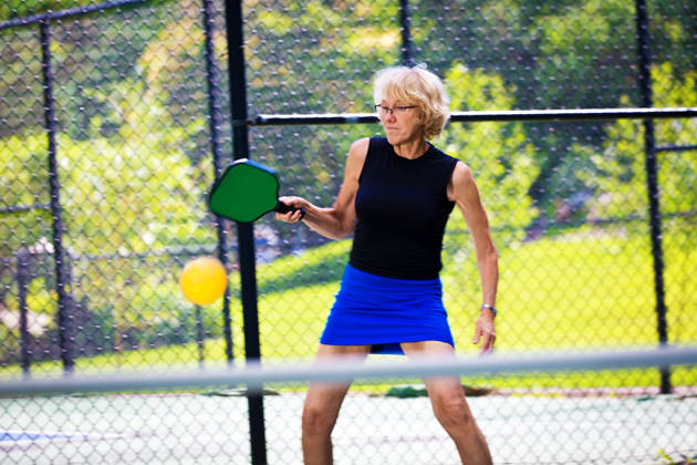 A Caucasian retiree adult woman pickleball player in action. She is holding a pickleball paddle posing to return the ball in a pickleball court. Photographed in horizontal format with copy space.
