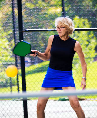 A Caucasian retiree adult woman pickleball player in action. She is holding a pickleball paddle posing to return the ball in a pickleball court. Photographed in horizontal format with copy space.
