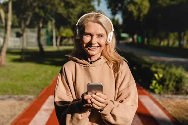 Cheerful sporty slim mature woman female runner athlete choosing sound track on smart phone while listening to the music radio podcast while training jogging in stadium
