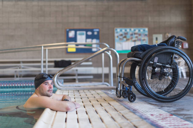 A paraplegic athlete finishes up his swim training in the pool. He is resting on the side of the pool by his wheelchair.
