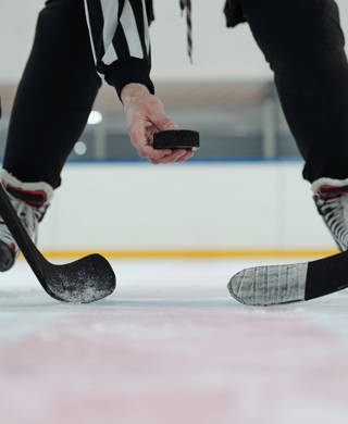 Hand of referee holding puck over ice rink with two players with sticks standing around and waiting for moment to shoot it